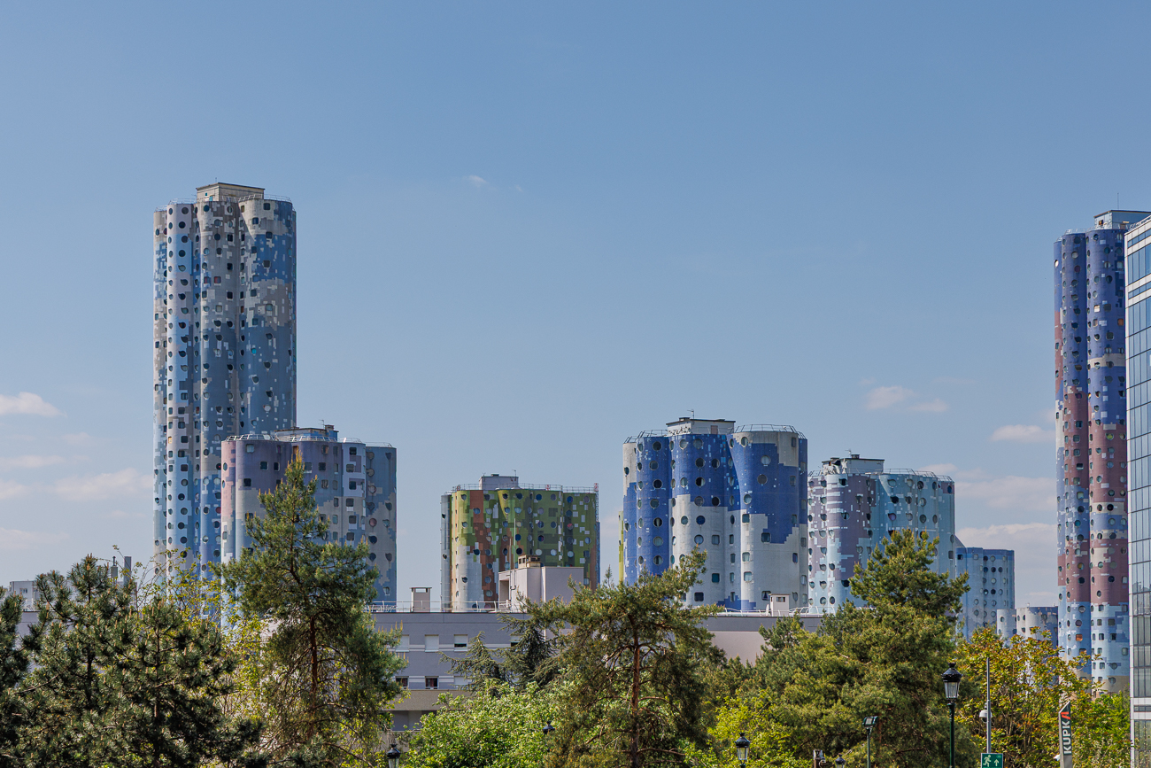 Die im Bild gezeigten Gebäude sind die Tours Aillaud, auch bekannt als Tours Nuages (Wolkentürme) oder Cité Pablo Picasso.Der Komplex besteht aus 18 Wohnhochhäusern mit charakteristischen zylinderförmigen Grundrissen und Fassaden, die Fresken von Wolken darstellen